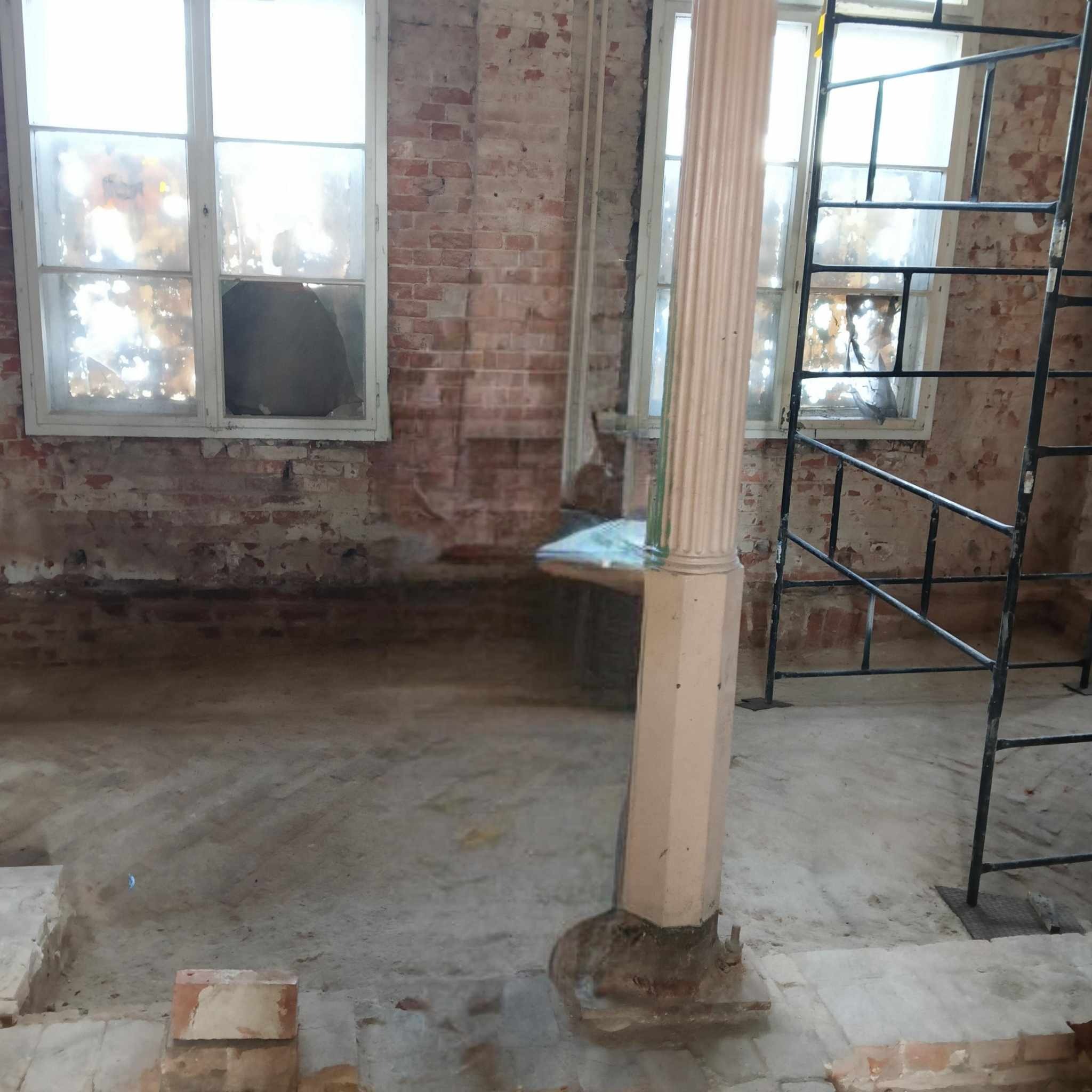 Wnętrze w trakcie remontu: odsłonięta cegła, okna z częściowo usuniętymi szybami, kolumna dekoracyjna zabezpieczona folią, rusztowanie, przygotowanie do dalszych prac wykończeniowych.