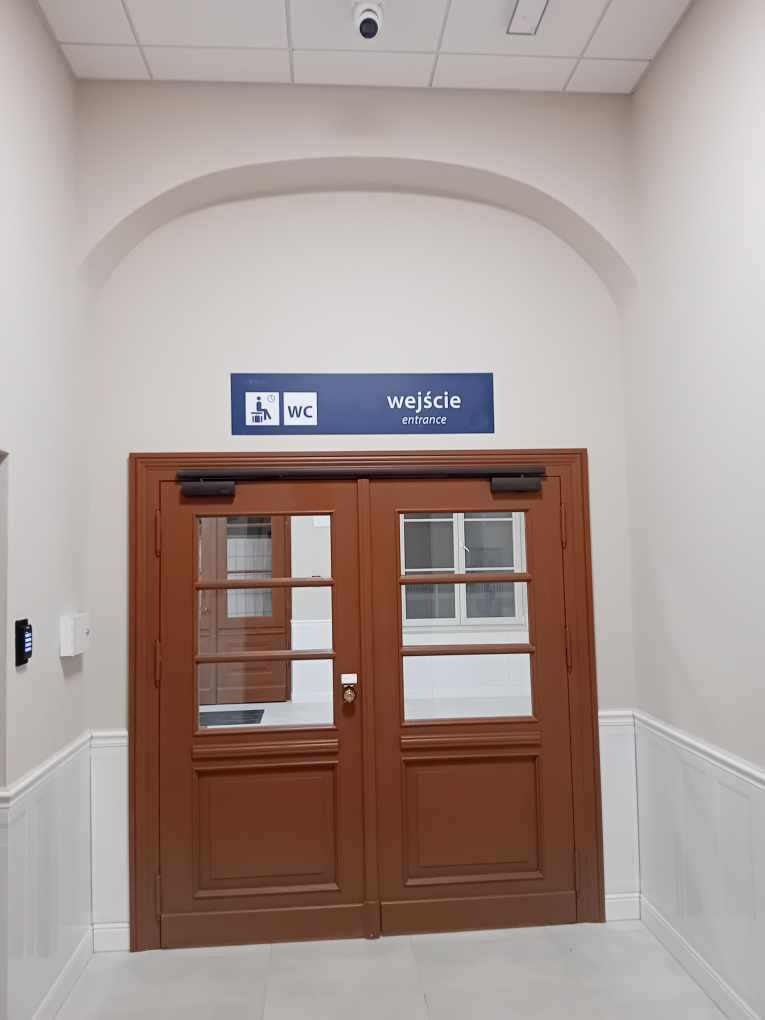 Brązowe, dwuskrzydłowe drzwi z przeszkleniami prowadzące do toalety, nad drzwiami tabliczka informacyjna z napisem 'wejście/entrance' i symbolem WC, widoczne elementy wykończenia ścian.