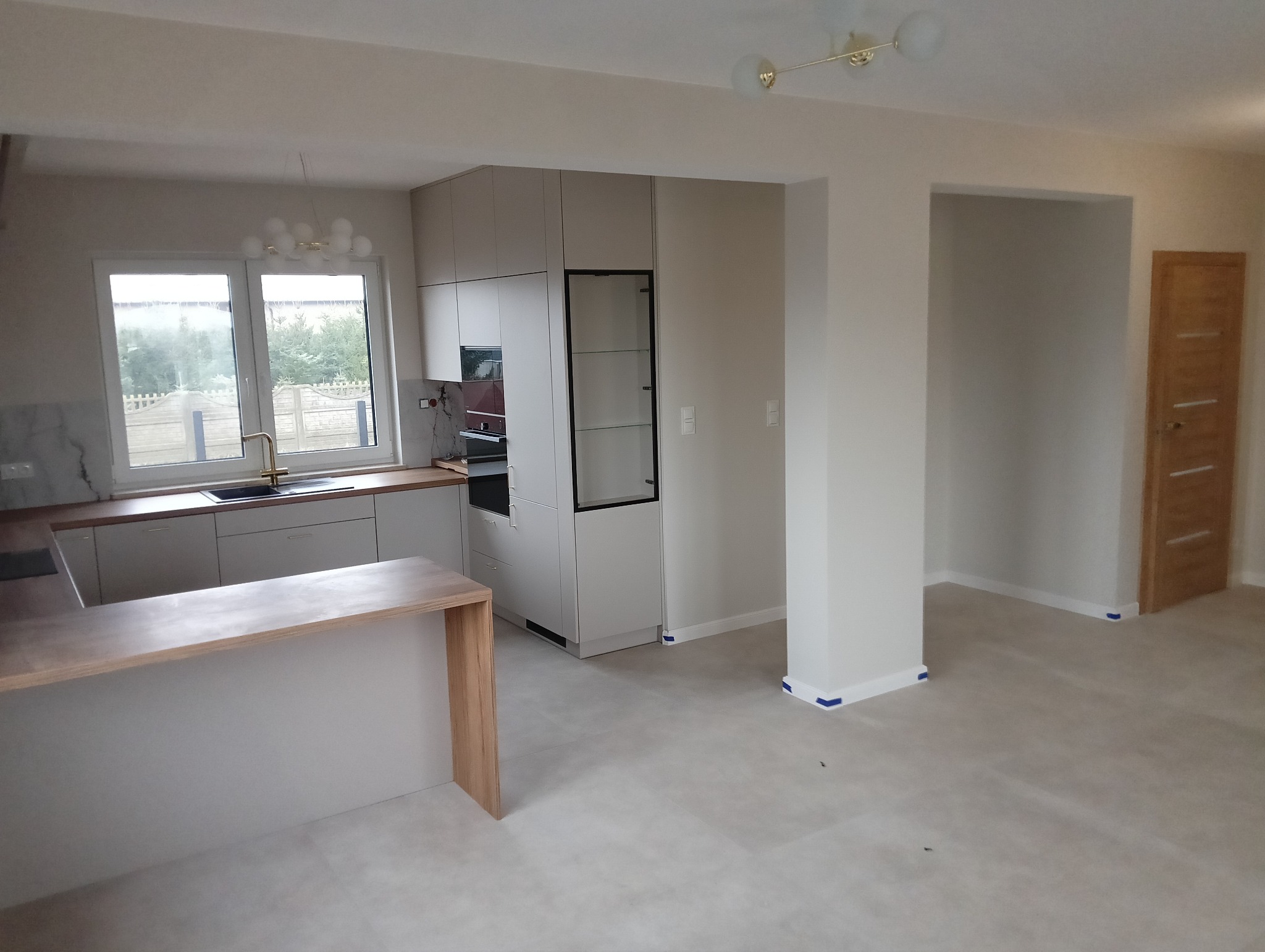 Jasne wnętrze po remoncie: widok na kuchnię z wyspą, świeżo pomalowane ściany i listwy przypodłogowe zabezpieczone taśmą malarską, drzwi wewnętrzne w tle.