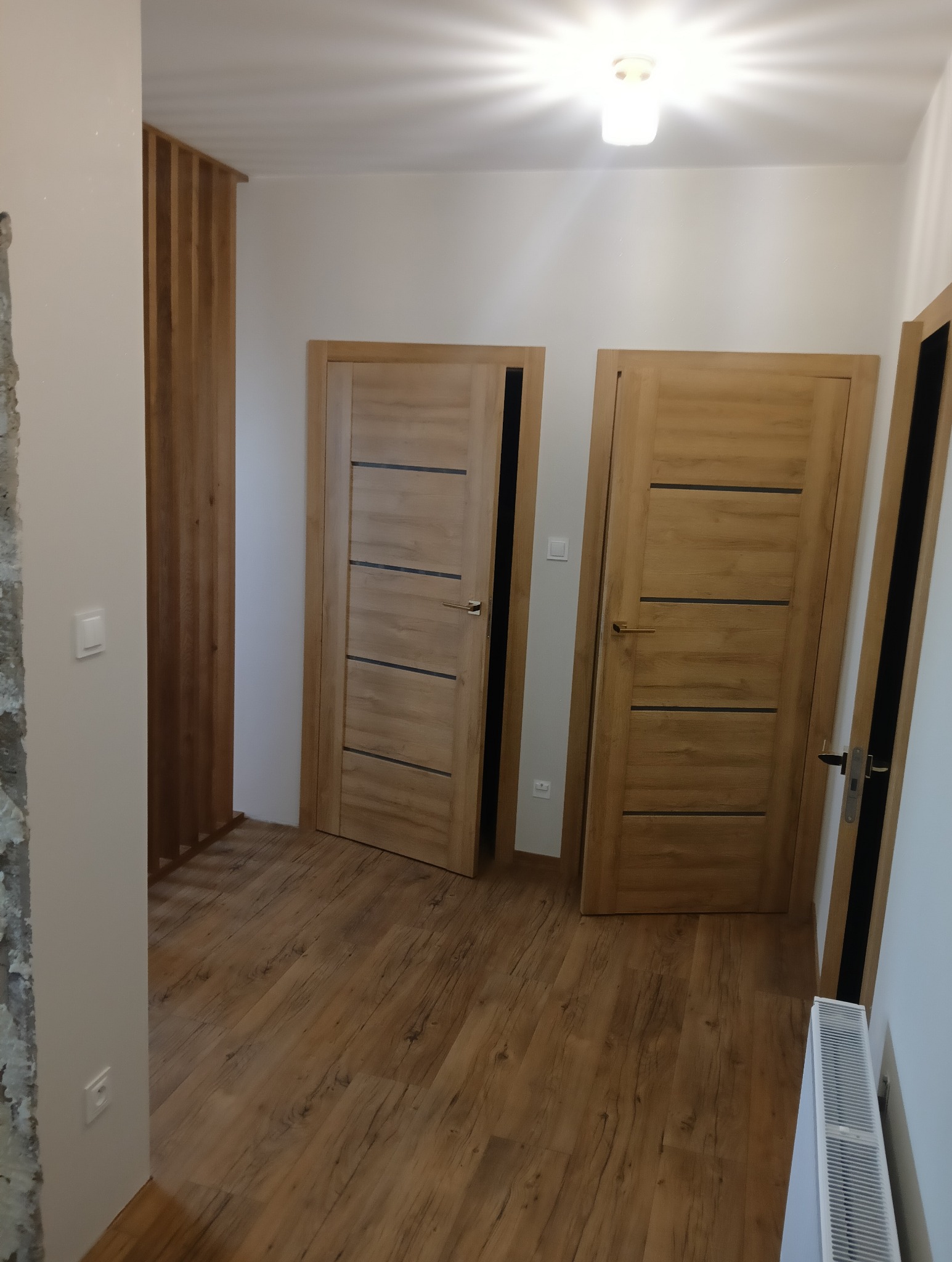 Wnętrze po remoncie: dwie drewniane drzwi z czarnymi akcentami, drewnopodobna podłoga, dekoracyjna ścianka z drewnianych listew, widoczny fragment ściany w trakcie wykończenia.