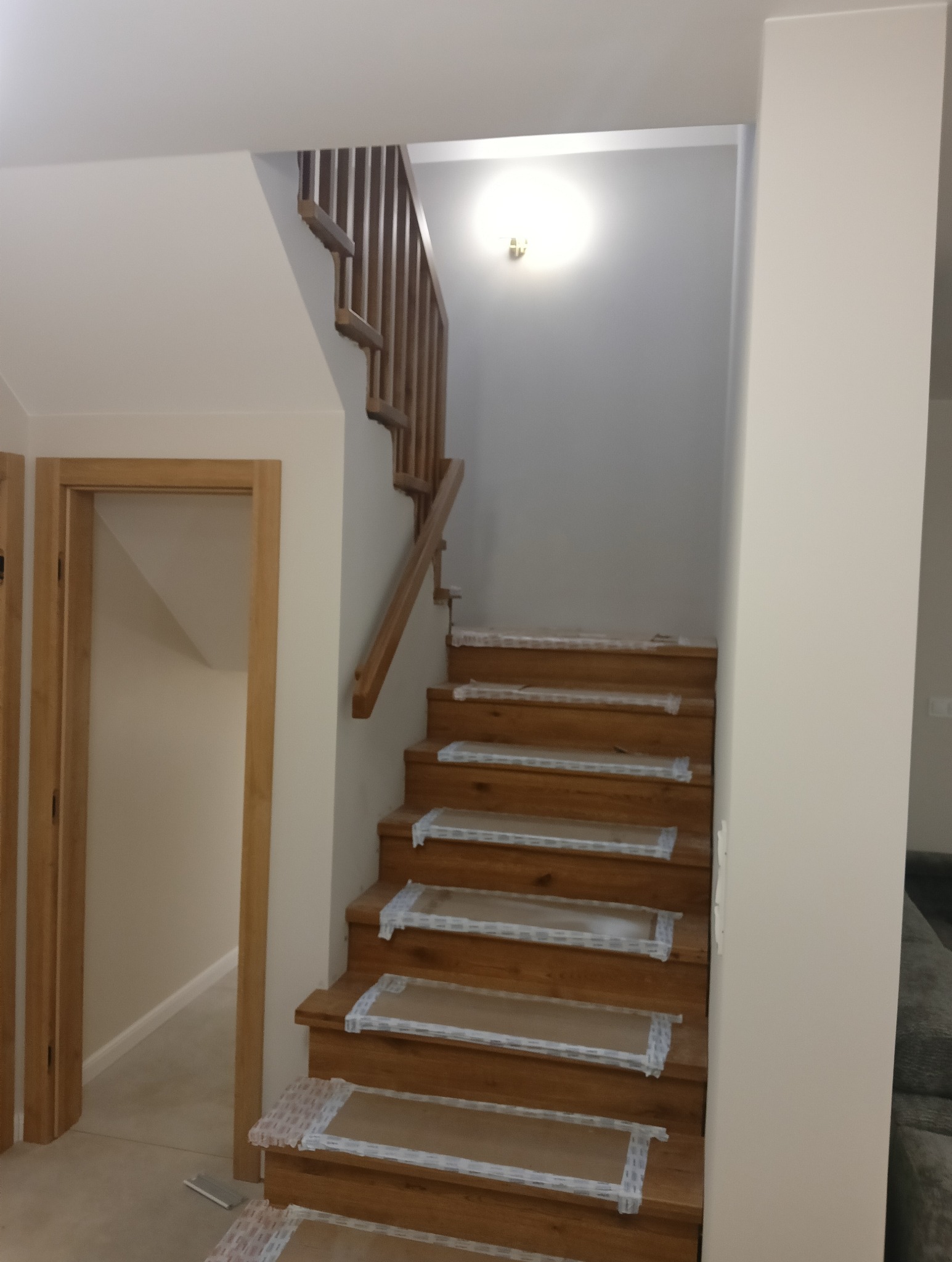 Drewniane schody z zabezpieczeniem na stopniach w trakcie remontu, widoczna drewniana balustrada i fragment pomalowanej ściany.