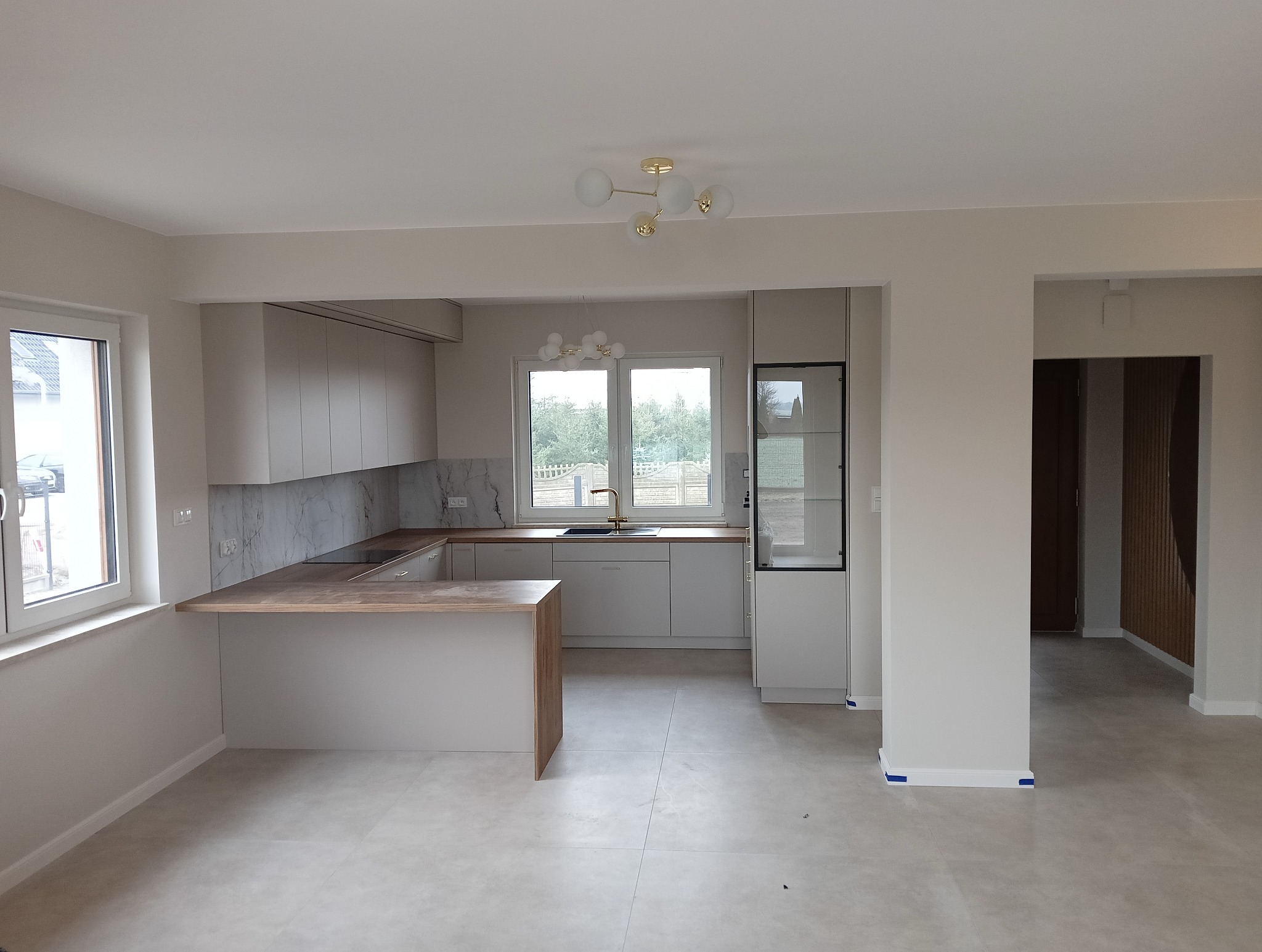 Jasna, nowoczesna kuchnia z wyspą, marmurowym backsplashiem i złotą armaturą, widok na salon z drewnianą ścianą.