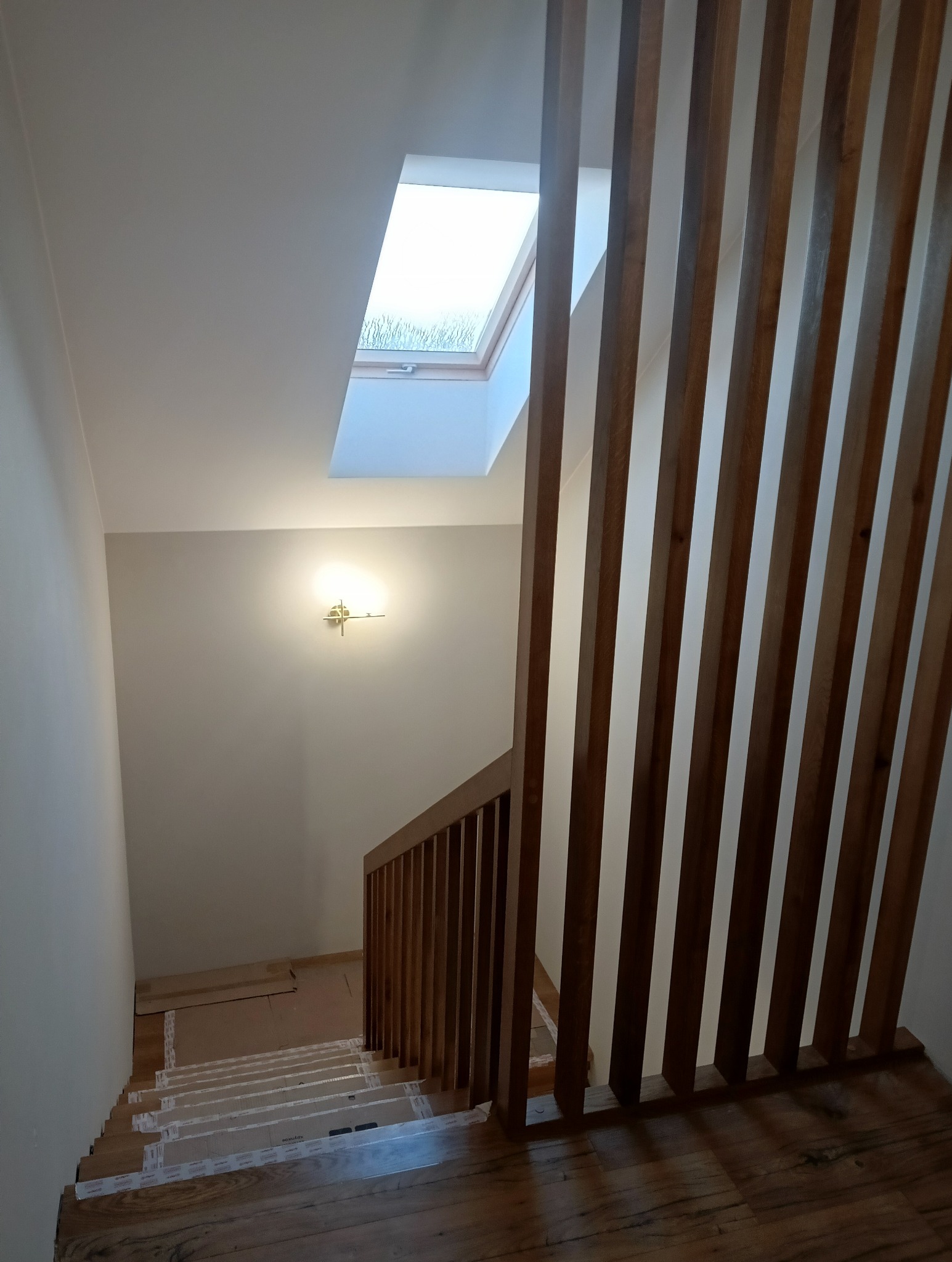 Schody drewniane z zabezpieczonymi stopniami, widok z góry, z pionowymi drewnianymi listwami balustrady i oknem dachowym w tle.