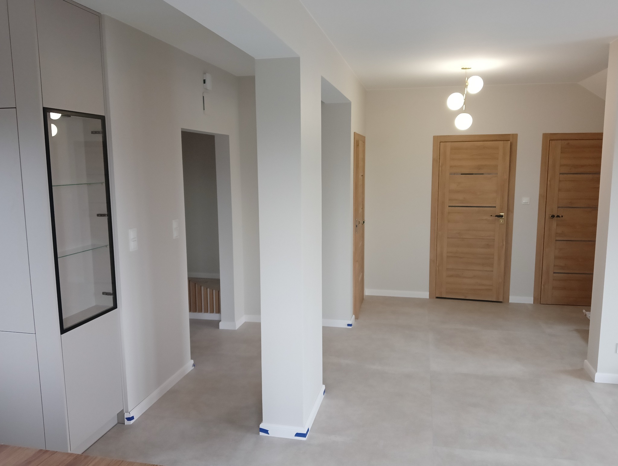 Jasne wnętrze po remoncie: szare płytki podłogowe, świeżo pomalowane ściany, dwoje drewnianych drzwi z poziomymi intarsjami, nowoczesna lampa sufitowa z kulistymi kloszami i biała zabudowa meblowa...