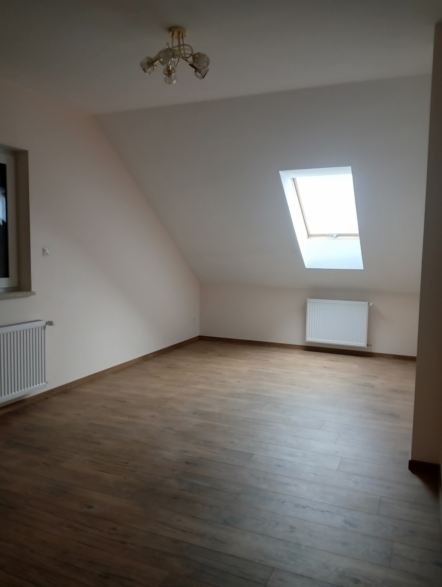 Poddasze po remoncie: Ściany w odcieniach beżu, panele podłogowe imitujące drewno, okno dachowe wpuszczające jasne światło, białe grzejniki i złoty żyrandol.