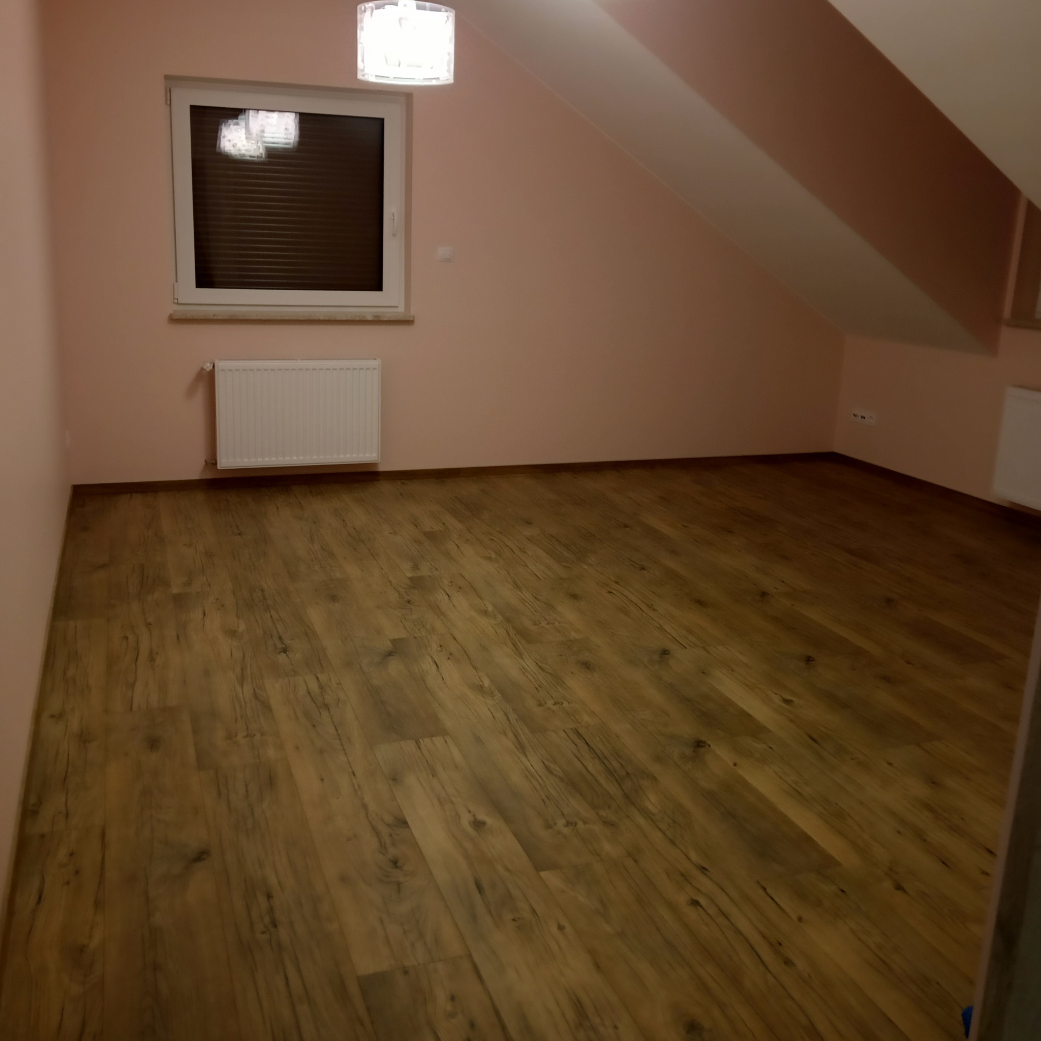 Pokój na poddaszu po remoncie: świeżo ułożone panele podłogowe w odcieniach brązu, ściany pomalowane na delikatny róż, okno z żaluzjami i biały grzejnik.