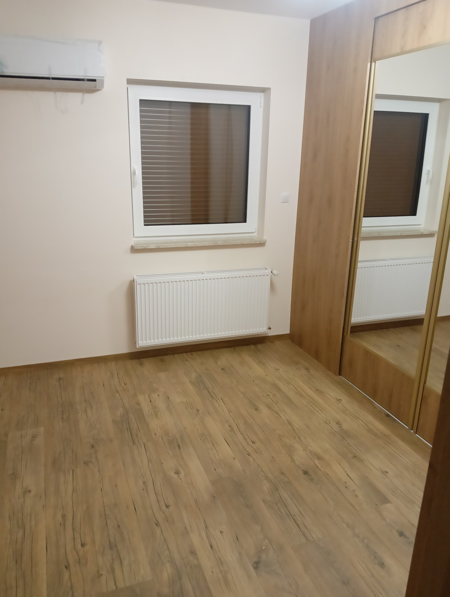 Puste wnętrze pokoju po remoncie z drewnopodobną podłogą, białym grzejnikiem, oknem z żaluzjami i szafą przesuwną z lustrem.