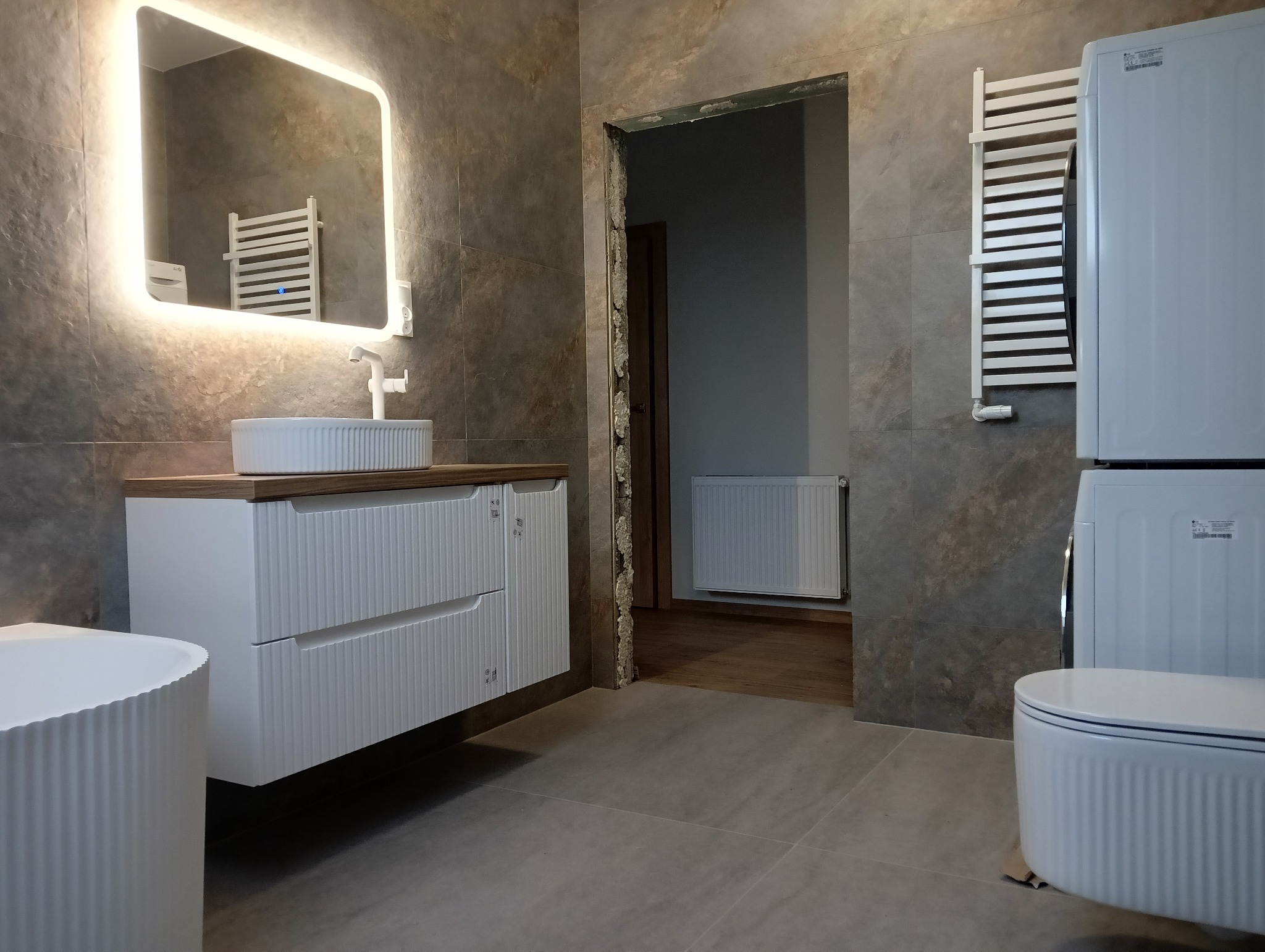 Nowoczesna łazienka z szarymi płytkami, podświetlanym lustrem nad białą szafką z umywalką, pralko-suszarką i grzejnikiem drabinkowym, widoczne niedokończone obramowanie drzwi.