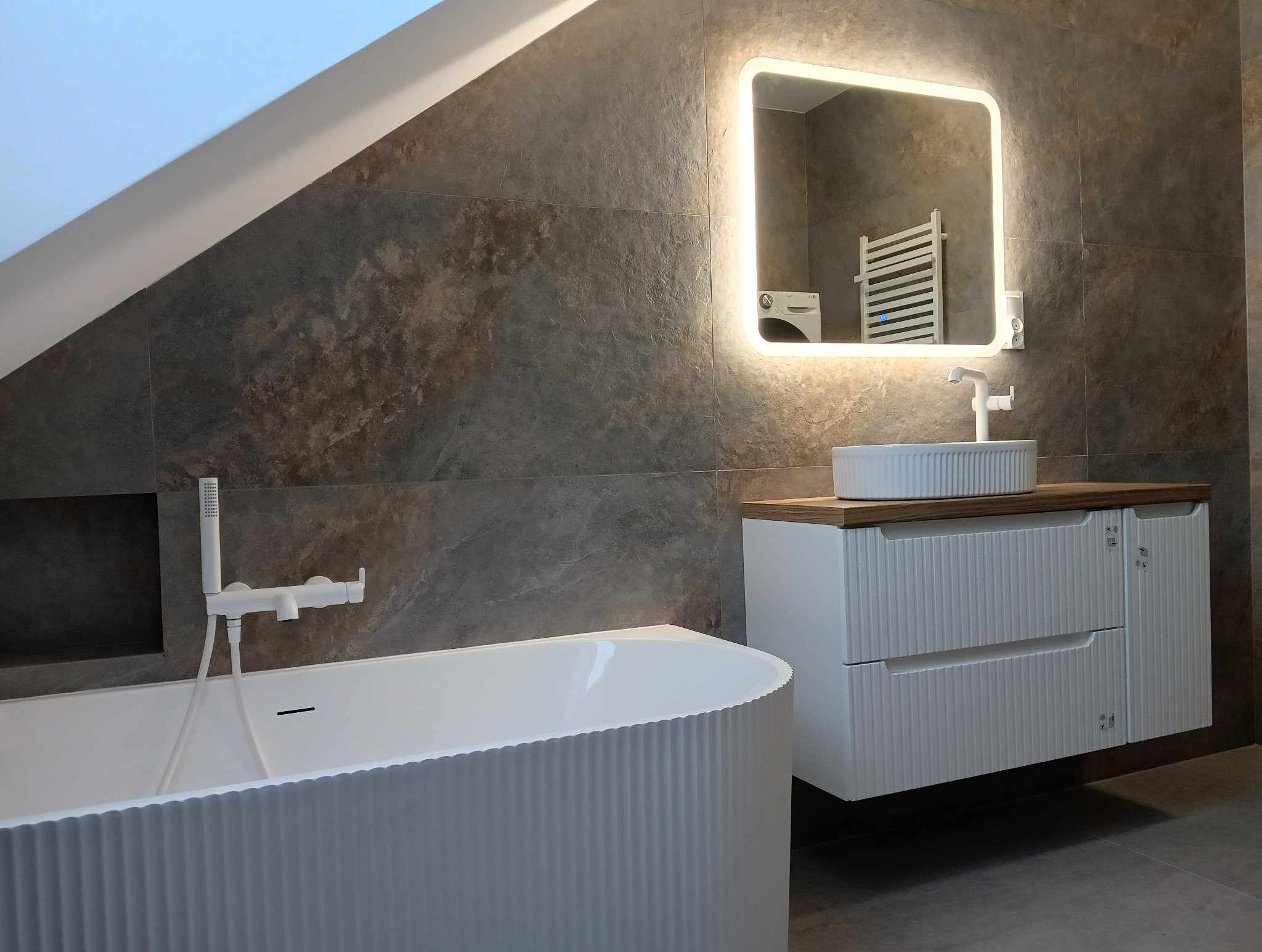 Nowoczesna łazienka z wanną o falistej strukturze, wiszącą szafką z umywalką i podświetlanym lustrem na tle płytek imitujących kamień.