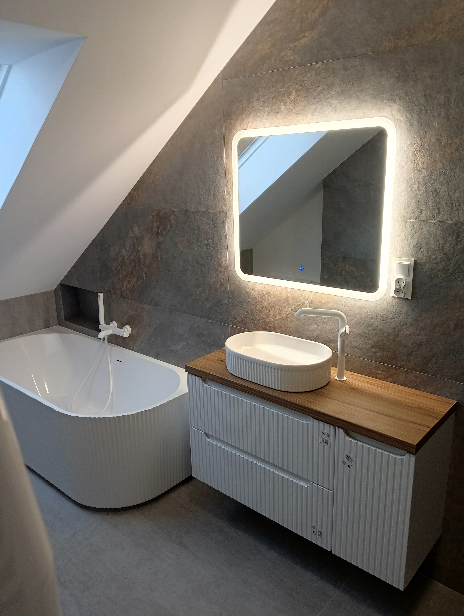 Nowoczesna łazienka na poddaszu z wanną o ryflowanej powierzchni, umywalką nablatową na drewnianym blacie i podświetlanym lustrem.