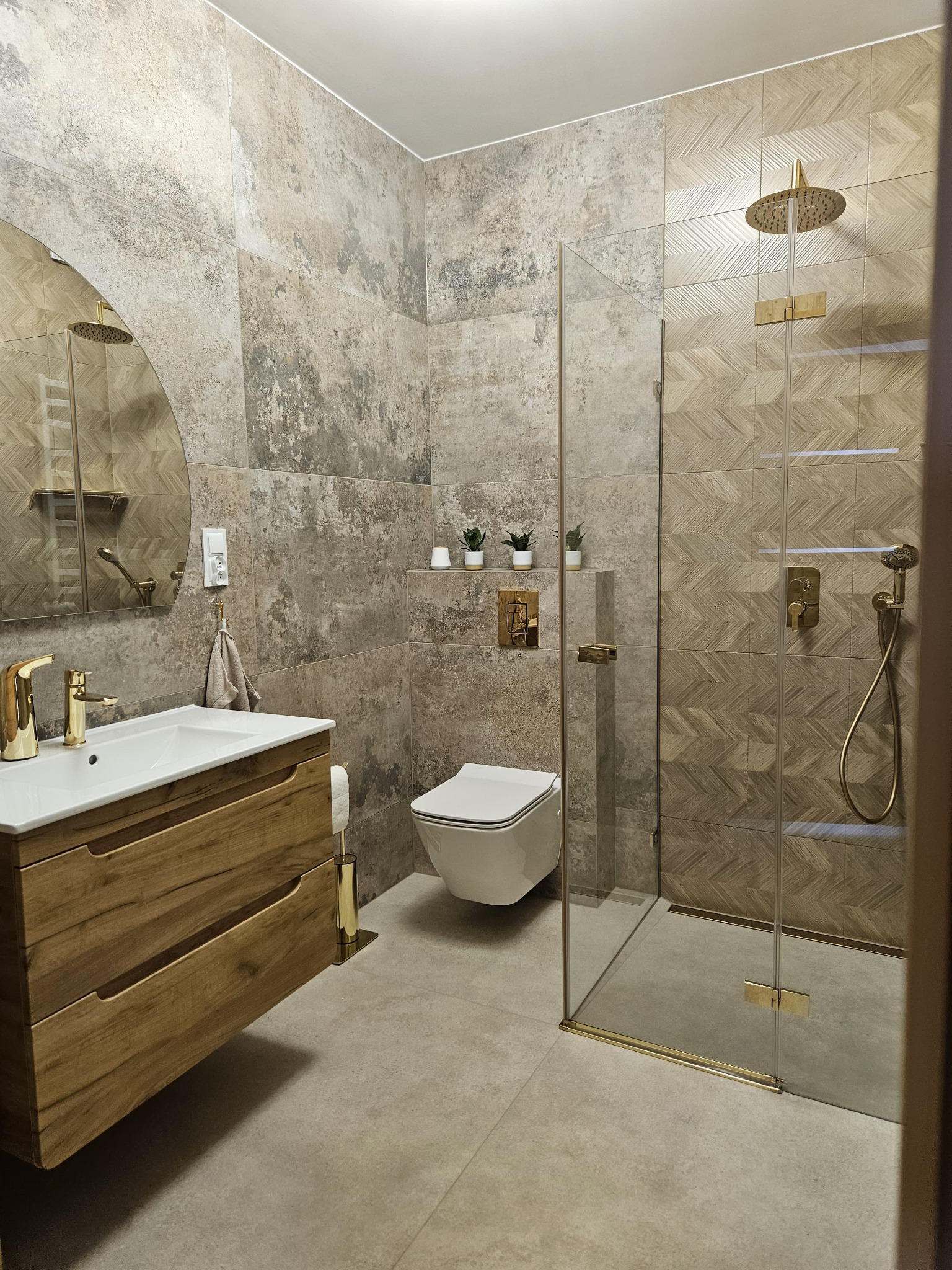 Nowoczesna łazienka z drewnianą szafką pod umywalką, złotą armaturą i kabiną prysznicową ze szklaną ścianką i wzorzystymi płytkami.