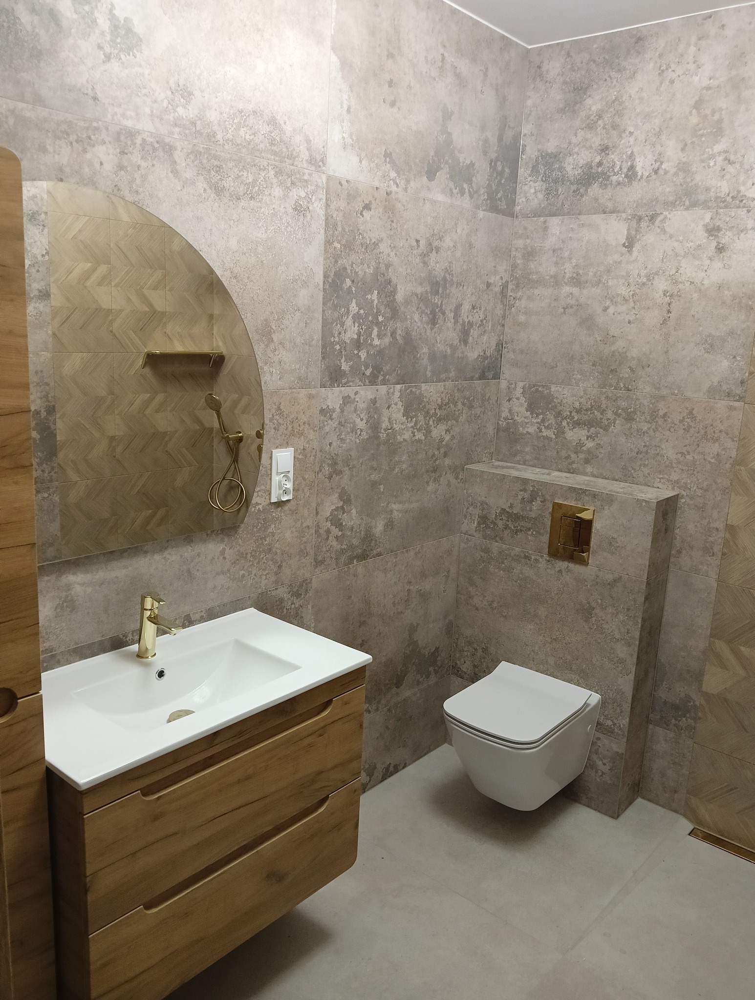 Nowoczesna łazienka z umywalką na drewnianej szafce, złotą armaturą, wiszącą toaletą i płytkami imitującymi beton na ścianach.
