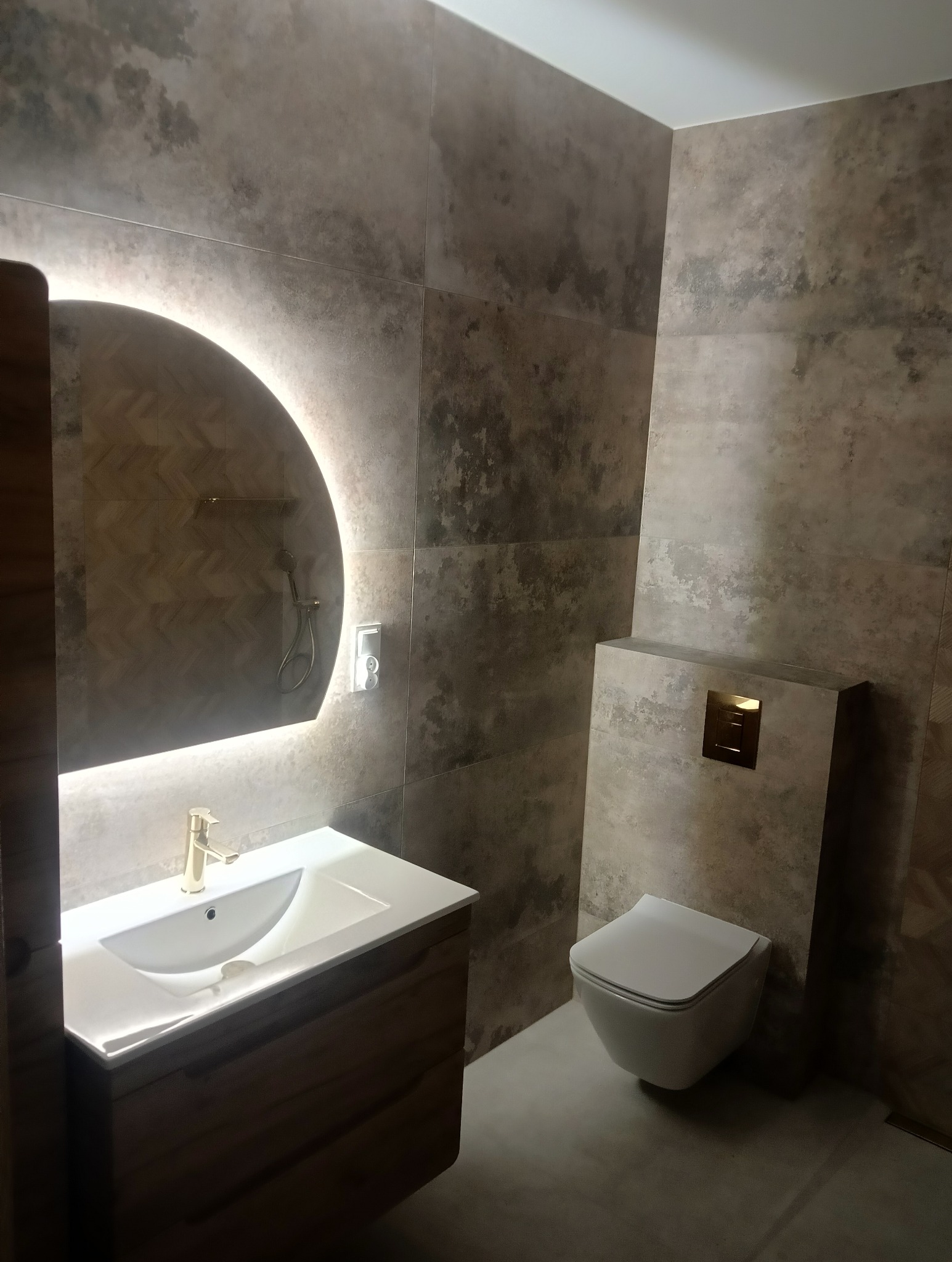 Nowoczesna łazienka z umywalką na drewnianej szafce, podświetlanym okrągłym lustrem i wiszącą toaletą, wykończona płytkami imitującymi beton.