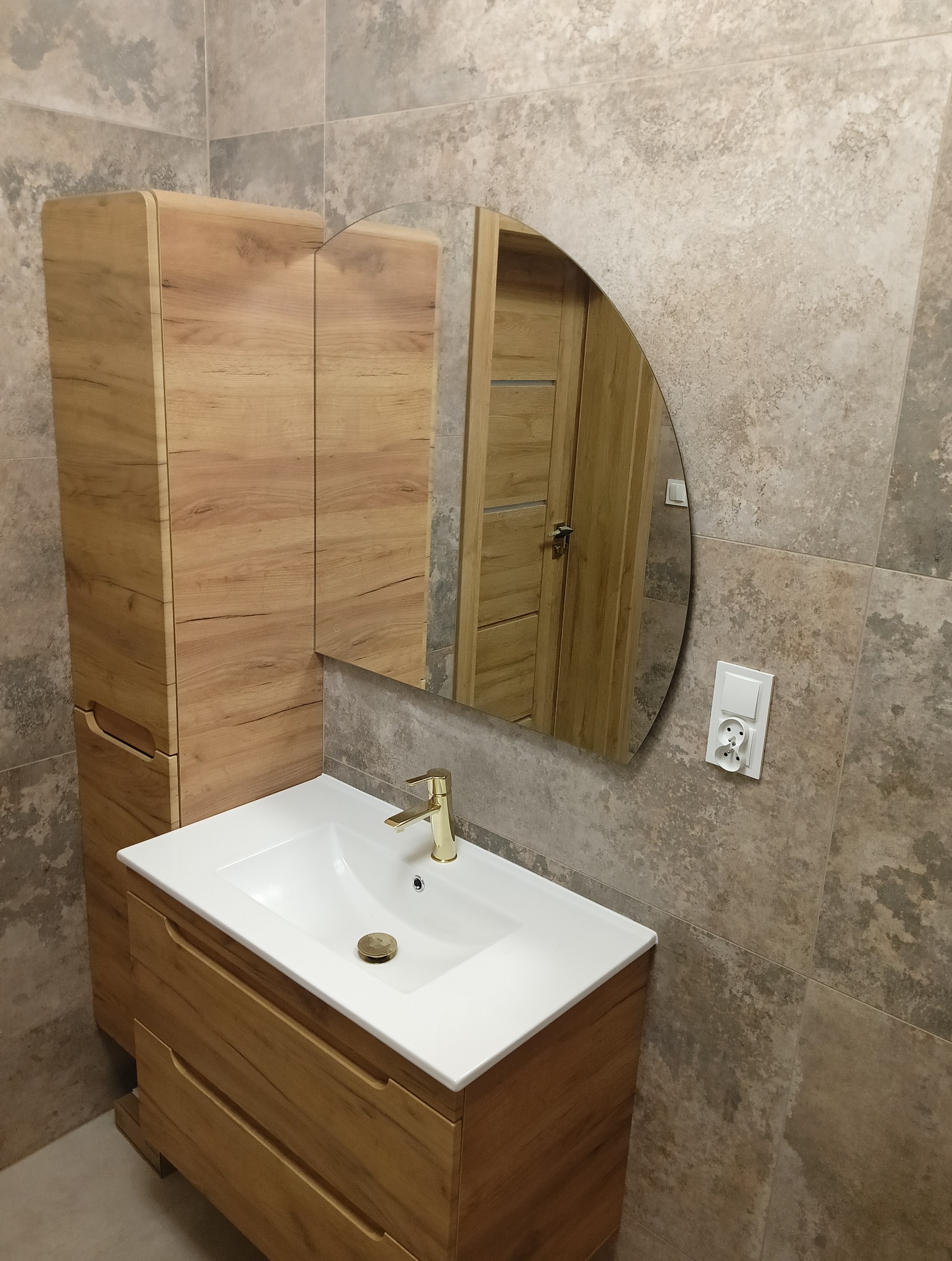Nowoczesna łazienka z drewnianą szafką pod umywalkę, owalnym lustrem i złotą baterią, wykończona płytkami w odcieniach beżu i szarości.