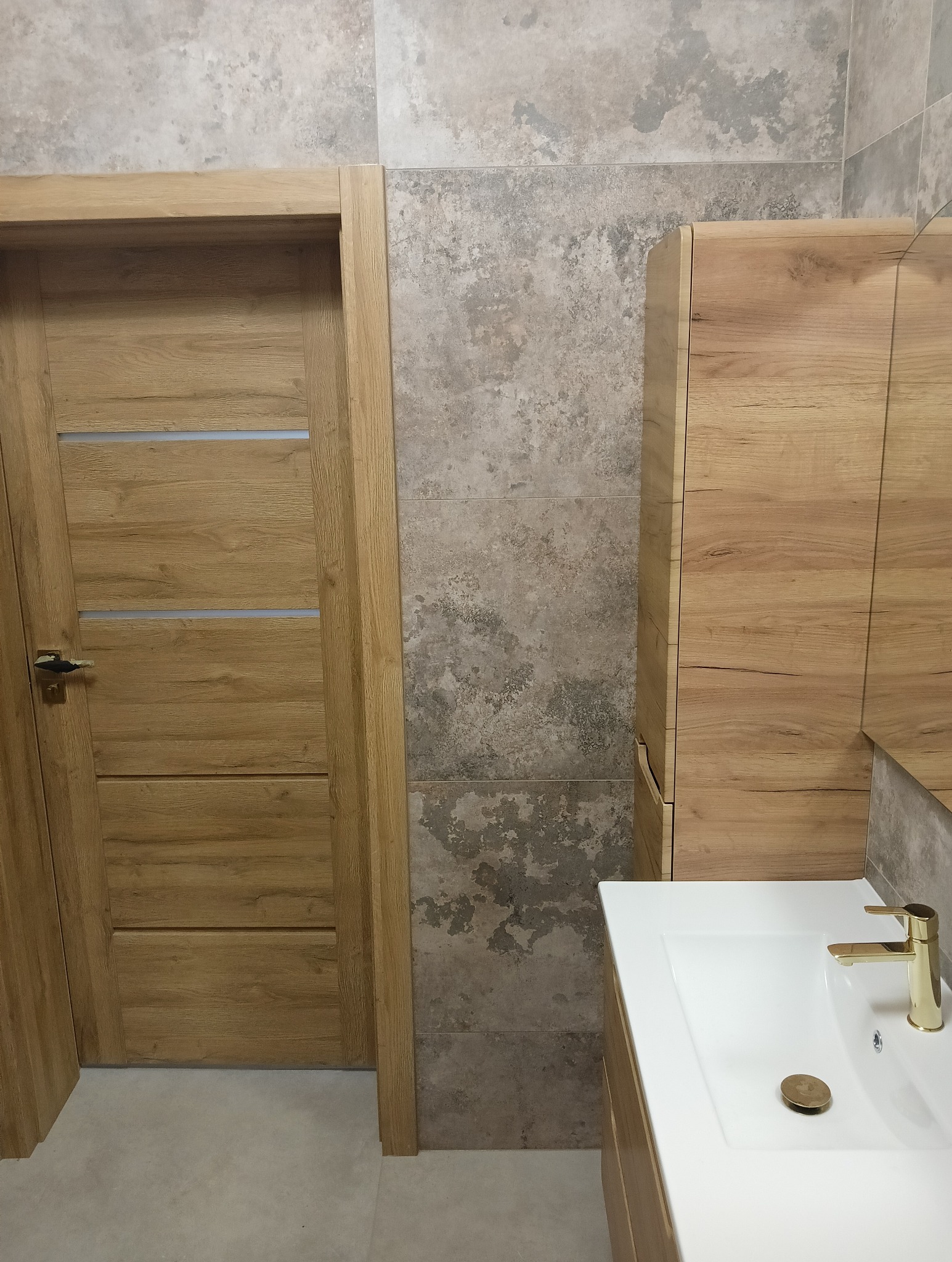 Nowoczesna łazienka z drewnianymi elementami: drzwi z poziomymi przeszkleniami, szafka pod umywalką i wysoka szafka wisząca, szara płytka imitująca kamień na ścianie.