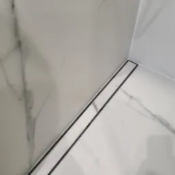 Biała łazienka wykończona płytkami imitującymi marmur z liniowym odpływem podłogowym z czarną ramką.