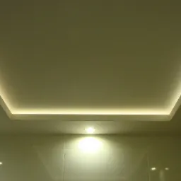 Podwieszany sufit z oświetleniem LED w kształcie prostokąta, dodatkowo mała lampka wbudowana w sufit, ściany wyłożone błyszczącymi płytkami w kolorze kremowym.