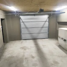 Wnętrze garażu po wylewce, z zamontowaną segmentową bramą garażową w kolorze białym, widoczne białe bloczki betonowe w rogu.