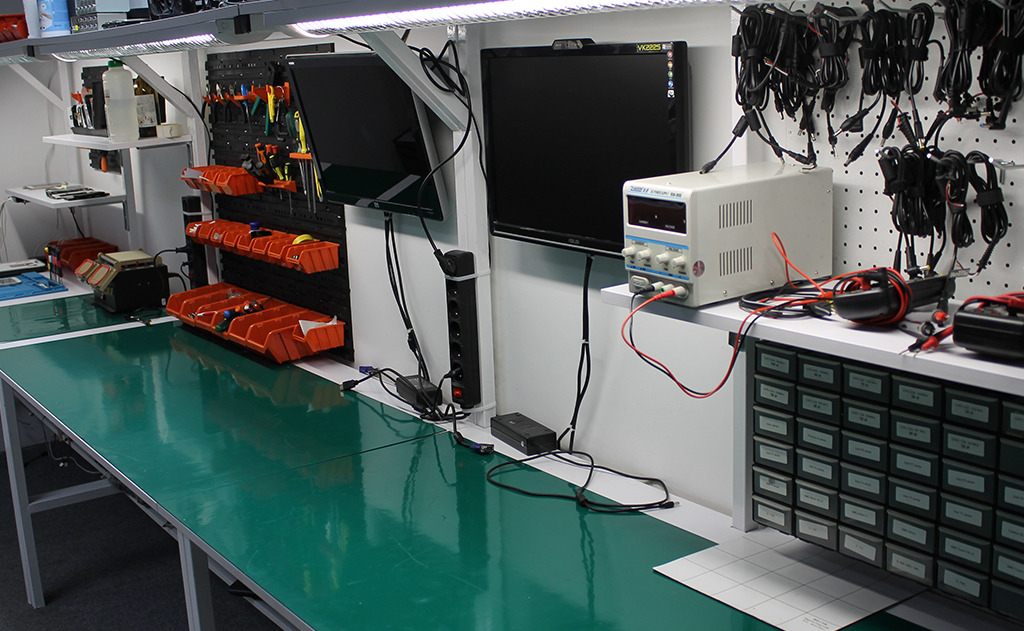 Stanowisko pracy z dwoma monitorami, zasilaczem laboratoryjnym, organizerami z narzędziami i zielonym blatem roboczym, oświetlone lampą LED.