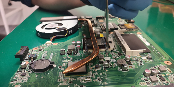 Demontaż układu chłodzenia laptopa: widoczna płyta główna z elementami, miedziana rurka cieplna, wentylator i ręka w niebieskiej rękawiczce używająca śrubokręta.