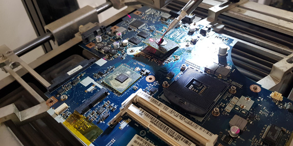 Płyta główna laptopa poddawana naprawie, precyzyjna aplikacja pasty termoprzewodzącej na układ scalony, widoczne elementy elektroniczne i uchwyt stabilizujący.