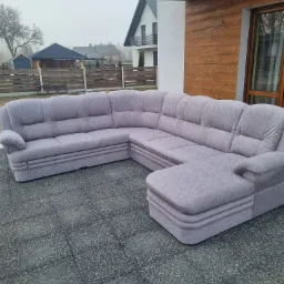 Szary narożnik modułowy z tapicerką na zewnątrz, ustawiony na szarych płytach chodnikowych przed nowoczesnym domem z drewnianą elewacją.