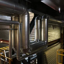 System rur wentylacyjnych ze stali nierdzewnej, z widocznymi łączeniami i elementami konstrukcyjnymi, umieszczony w przestrzeni przemysłowej z żółtymi barierkami.