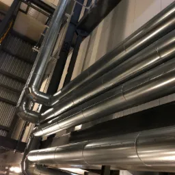 Instalacja przemysłowa: szereg równoległych, metalowych rur wentylacyjnych, widoczne łączenia segmentów, mocowania do konstrukcji stalowej pod sufitem hali.