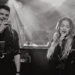 Czarno-białe zdjęcie duetu wokalnego podczas występu na scenie, mężczyzna z mikrofonem trzyma tablet na specjalnym uchwycie, kobieta śpiewa z zamkniętymi oczami, w tle sprzęt nagłośnieniowy...