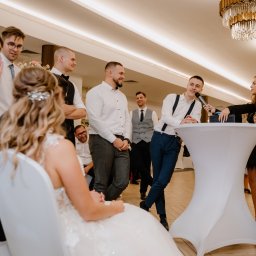 Zespół LepiNieBydzie - Mężczyzna w szelkach przemawia do mikrofonu podczas wesela, otoczony przez gości, z panną młodą siedzącą na pierwszym planie i kryształowym żyrandolem w tle.