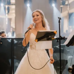 Uśmiechnięta piosenkarka w białej sukni śpiewa do mikrofonu na scenie, za nią widoczny fragment zespołu i oświetlenie sceniczne.