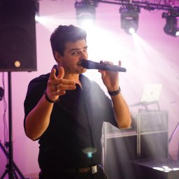 Zespół LepiNieBydzie - Wokalista w czarnej koszuli śpiewa do mikrofonu na scenie oświetlonej fioletowymi światłami, z widocznymi głośnikami i sprzętem nagłośnieniowym w tle.