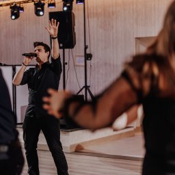 Zespół LepiNieBydzie - Wokalista w czarnym stroju i czerwonych trampkach śpiewa do mikrofonu na scenie, z podniesioną ręką, oświetlenie sceniczne w tle.