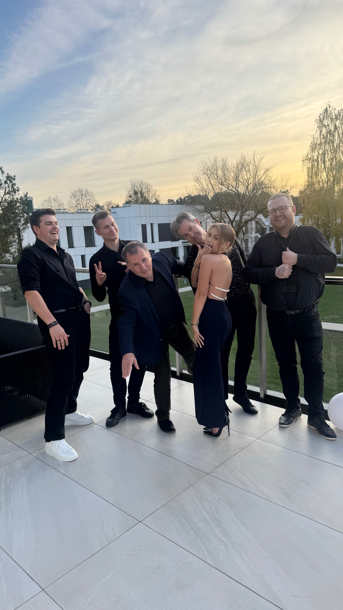 Uśmiechnięta grupa pięciu mężczyzn w czarnych strojach i kobiety w granatowej sukni pozuje na balkonie z szarą podłogą na tle budynku i zachodzącego słońca.