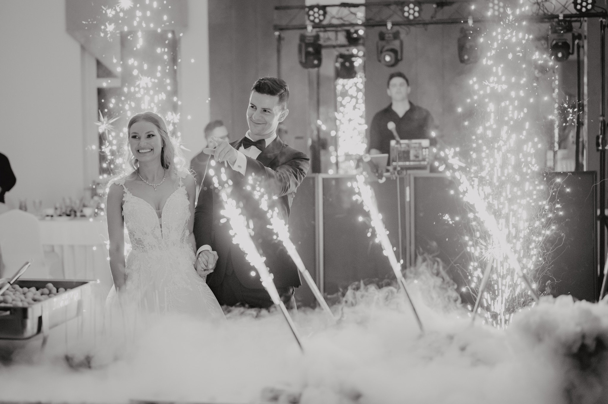 Para młoda w trakcie pierwszego tańca, otoczona fontannami iskier i dymem scenicznym; w tle DJ na stanowisku.