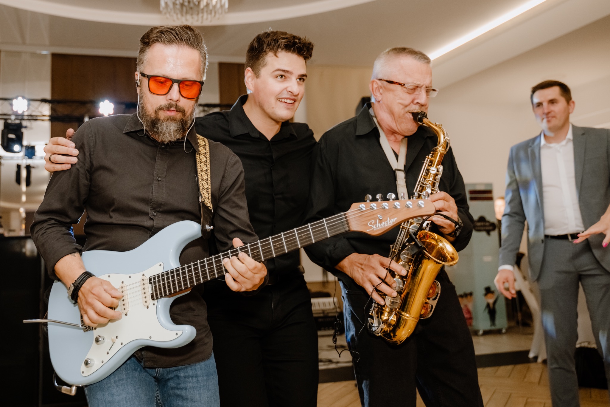 Trzech muzyków w Rychwale grających na gitarze elektrycznej (Schecter) i saksofonie podczas występu na imprezie; jeden z muzyków ma czerwone okulary.