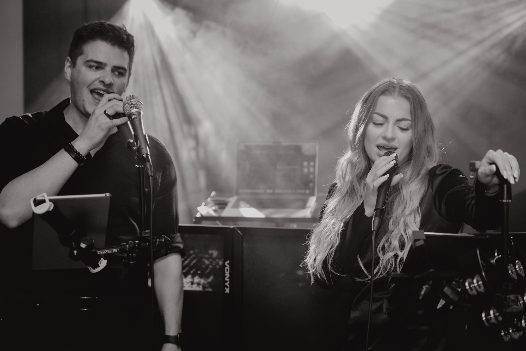 Czarno-białe zdjęcie duetu wokalnego podczas występu na scenie, mężczyzna z mikrofonem trzyma tablet na specjalnym uchwycie, kobieta śpiewa z zamkniętymi oczami, w tle sprzęt nagłośnieniowy...