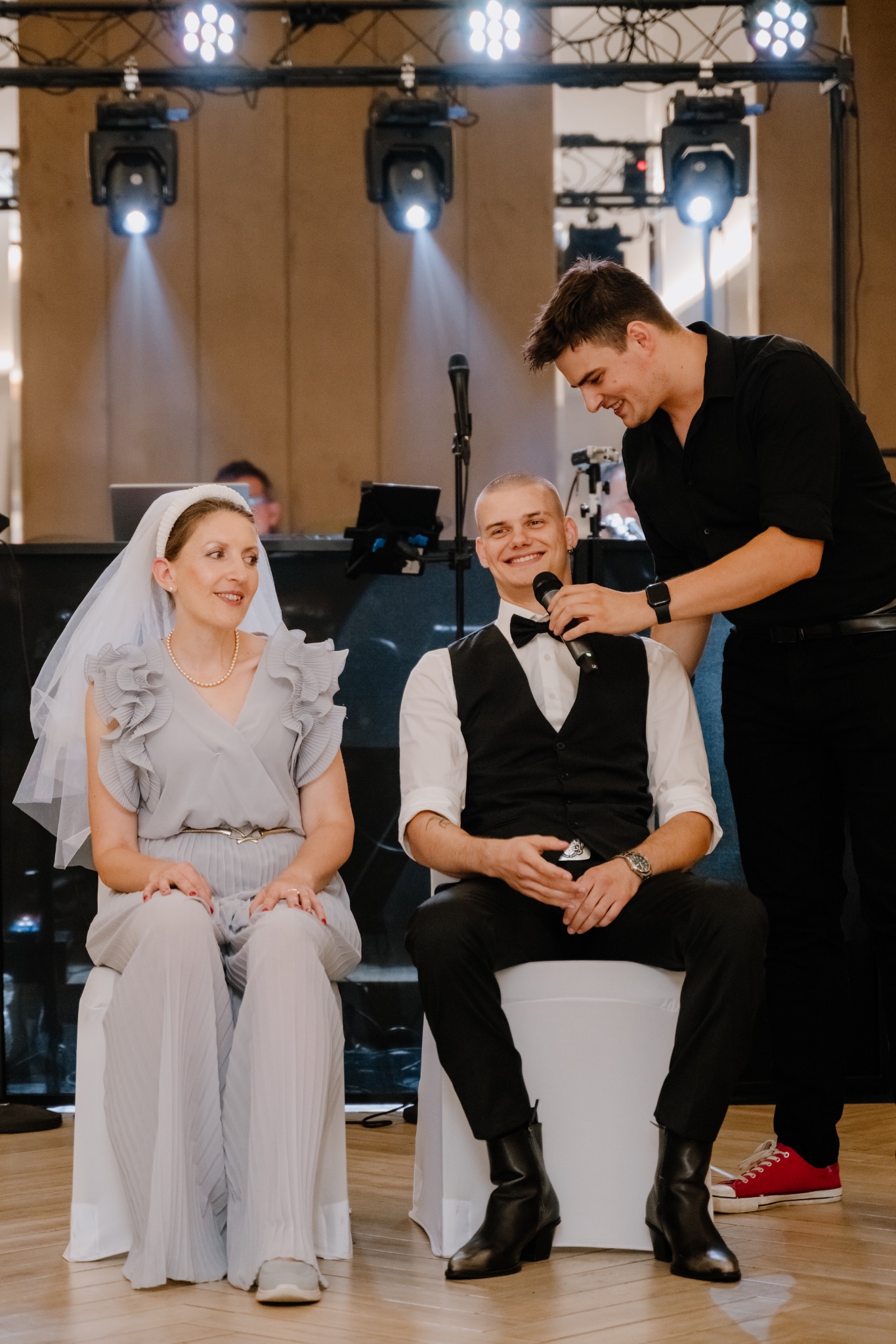 Panna młoda w szarej sukience i welonie siedzi obok pana młodego w muszce, podczas gdy mężczyzna w czarnej koszuli trzyma mikrofon przy jego ustach, oświetlenie sceniczne w tle.
