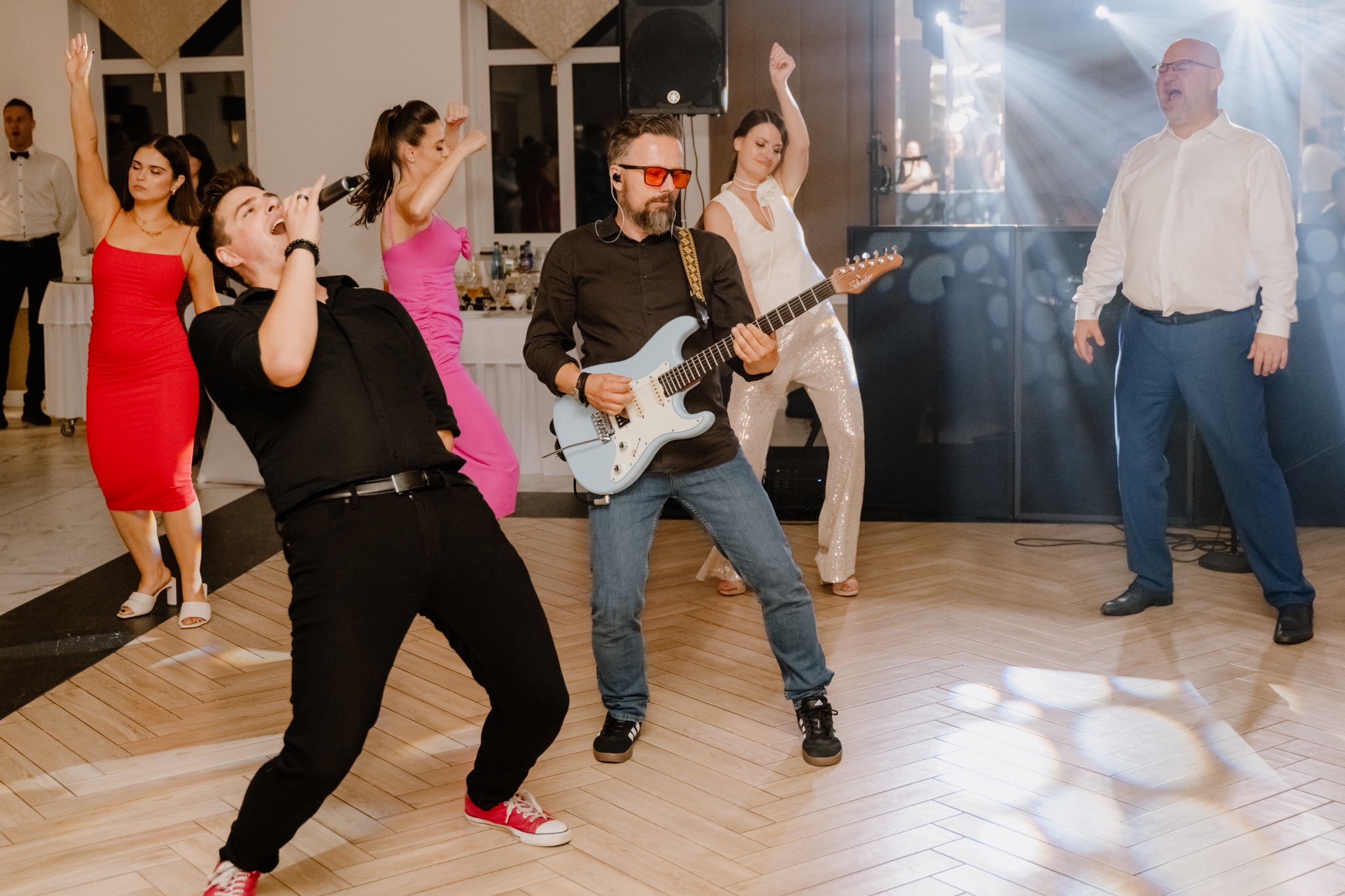 Wokalista w czarnej koszuli i spodniach, z czerwonymi trampkami, śpiewa z mikrofonem na scenie, obok gitarzysta z niebieską gitarą, w tle tańczące kobiety w sukienkach i mężczyzna w białej koszuli.