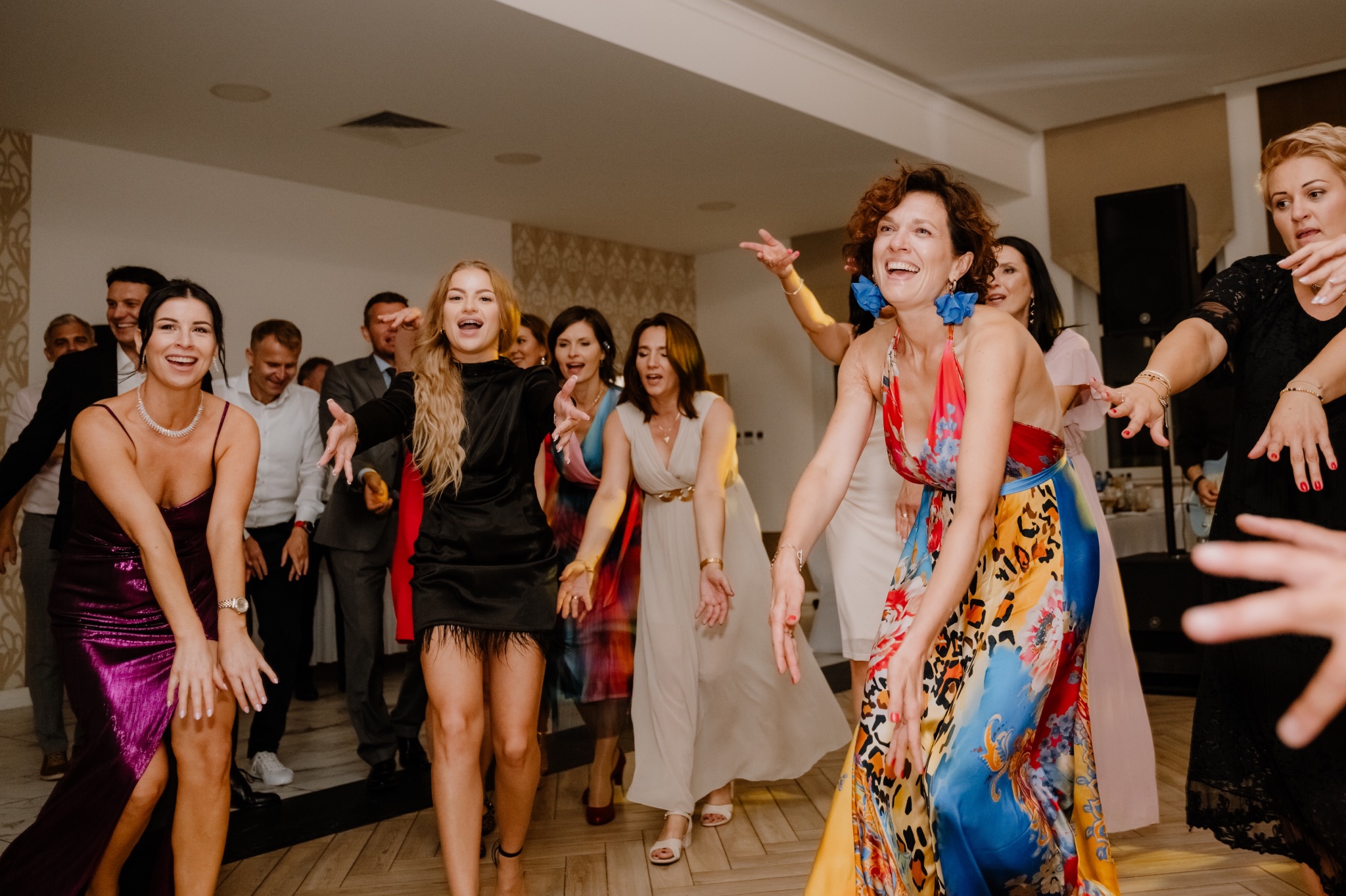 Rozbawieni goście weselni w kolorowych sukienkach tańczą na parkiecie podczas uroczystości, w tle widoczni inni uczestnicy zabawy.