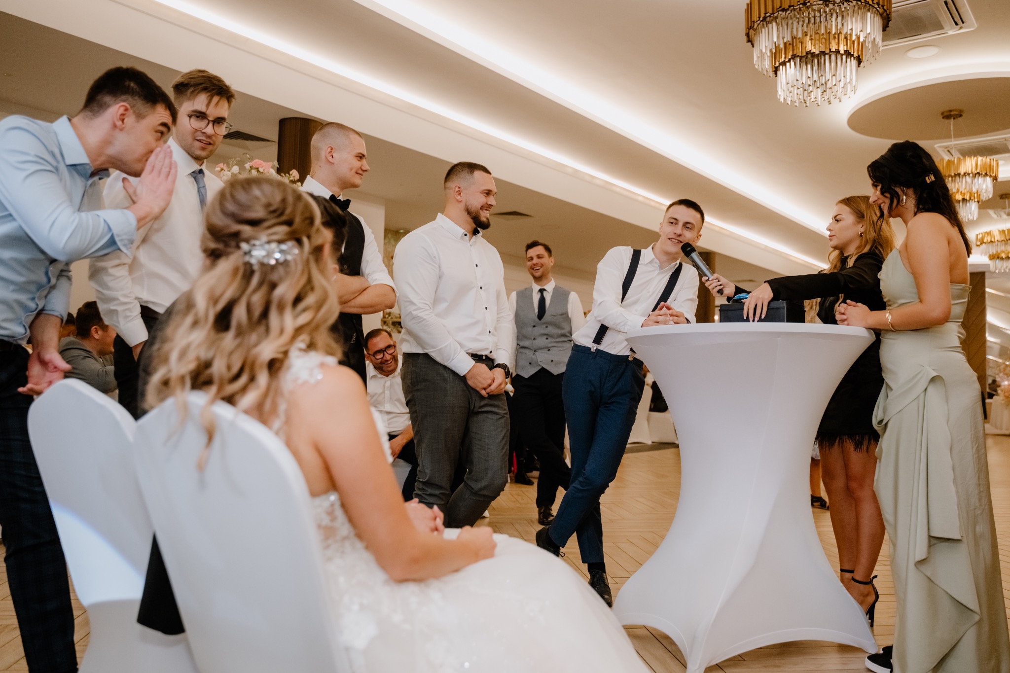 Mężczyzna w szelkach przemawia do mikrofonu podczas wesela, otoczony przez gości, z panną młodą siedzącą na pierwszym planie i kryształowym żyrandolem w tle.