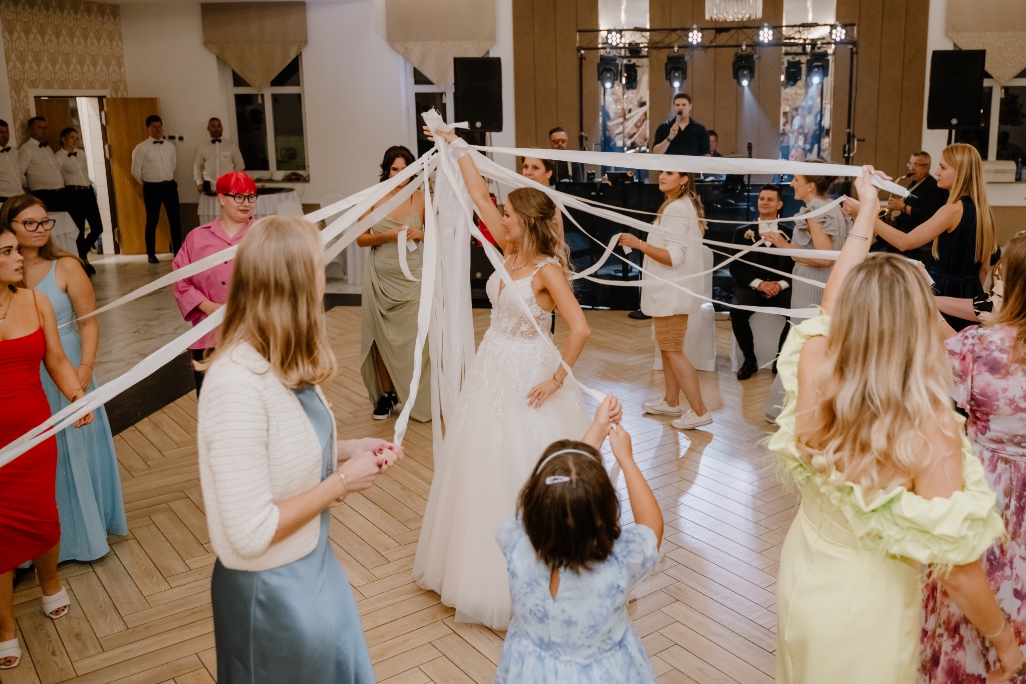 Panna młoda w białej sukni tanecznej otoczona gośćmi trzymającymi białe serpentyny podczas zabawy oczepinowej, na scenie w tle widać zespół muzyczny z mikrofonem i saksofonem.