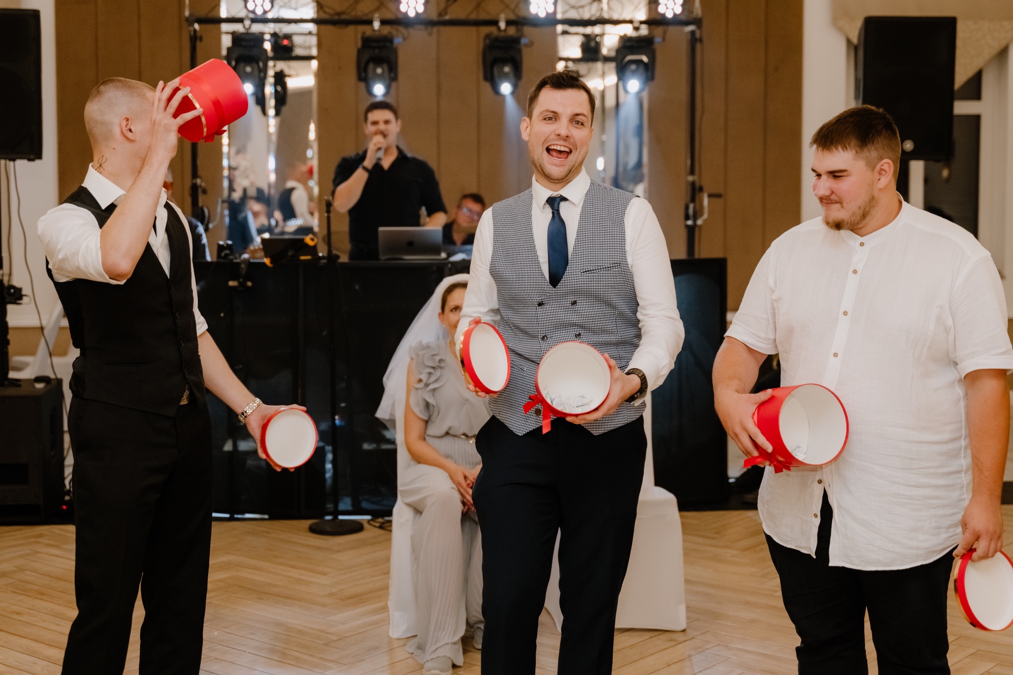 Trzech mężczyzn w eleganckich strojach trzyma otwarte czerwone pudełka na tle sceny z DJ-em i parą młodą w tle, podczas uroczystości.