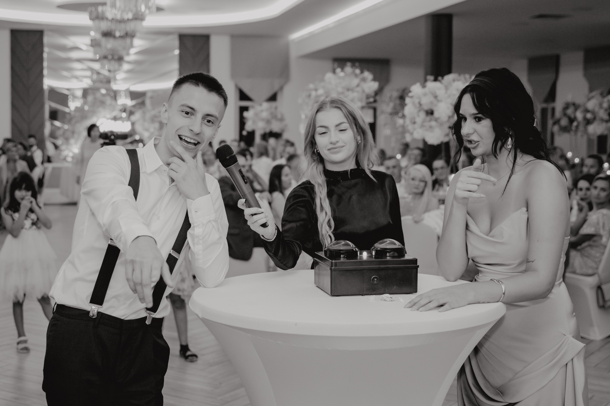 Czarno-białe zdjęcie konferansjera z mikrofonem, kobiety z urządzeniem do głosowania oraz kobiety w sukni wieczorowej, prawdopodobnie podczas konkursu na weselu.