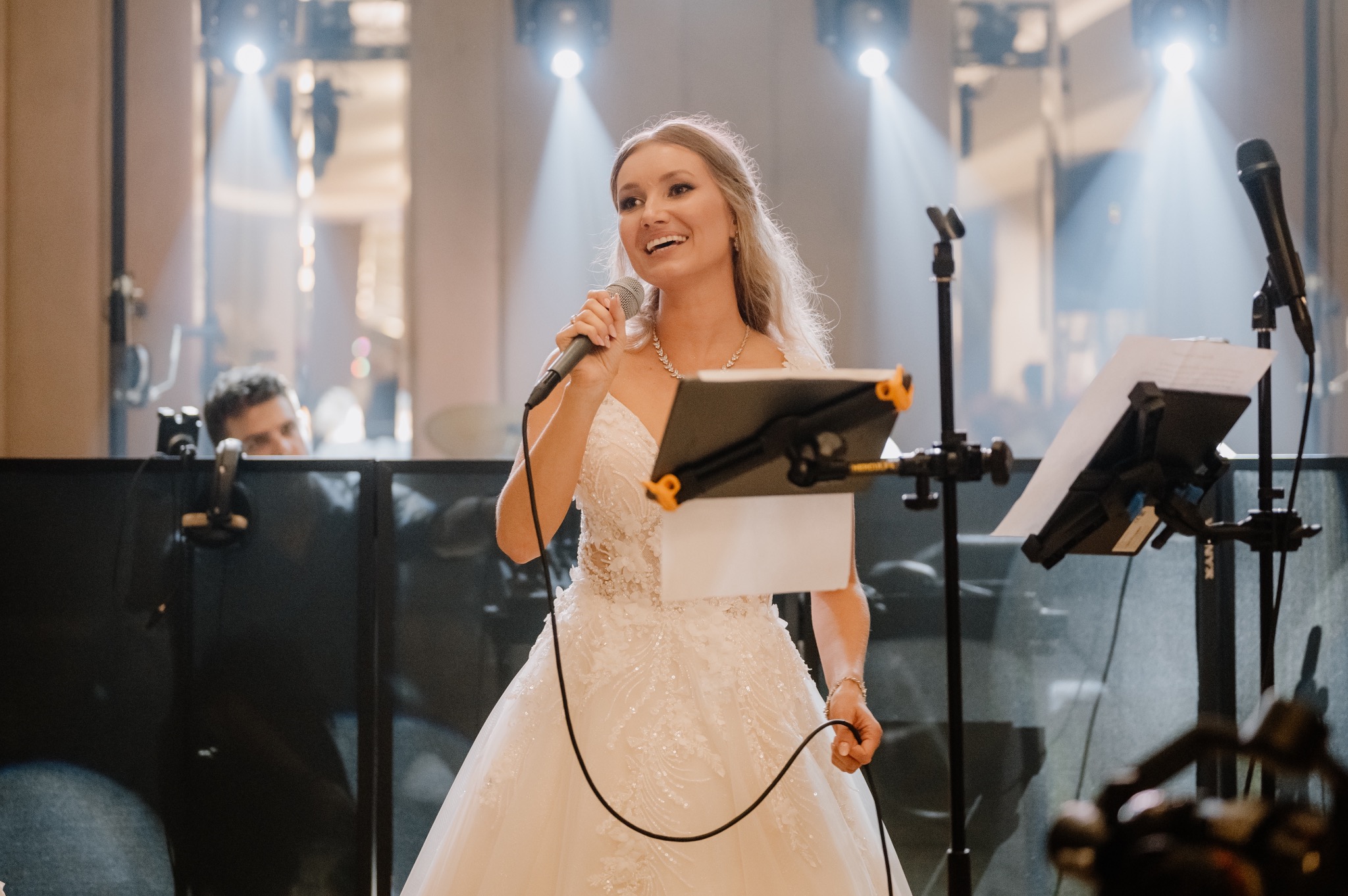 Uśmiechnięta piosenkarka w białej sukni śpiewa do mikrofonu na scenie, za nią widoczny fragment zespołu i oświetlenie sceniczne.