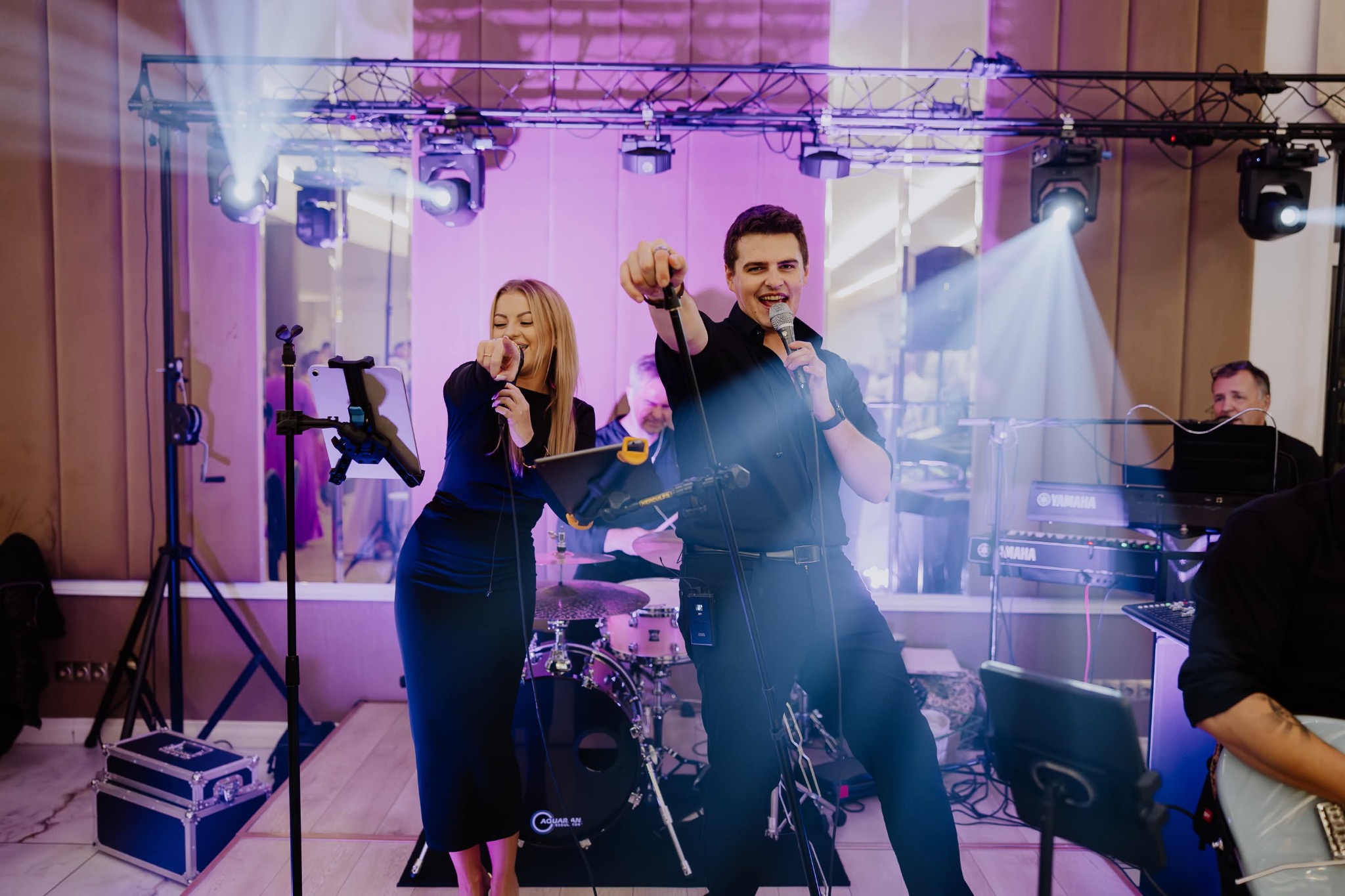 Zespół muzyczny w akcji: wokalistka w długiej sukni i wokalista z mikrofonem, perkusja, keyboard Yamaha, oświetlenie sceniczne w fioletowych barwach, dynamiczne ujęcie.