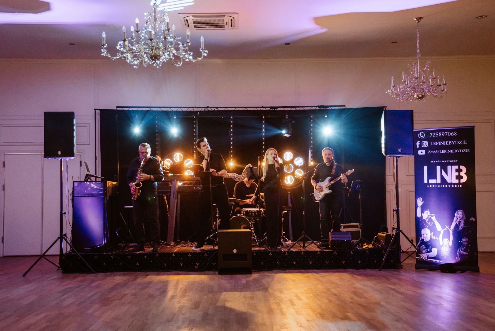 Zespół muzyczny na scenie podczas występu w sali balowej z żyrandolami. Saksofonista, wokaliści, perkusista i gitarzysta w czarnych strojach.
