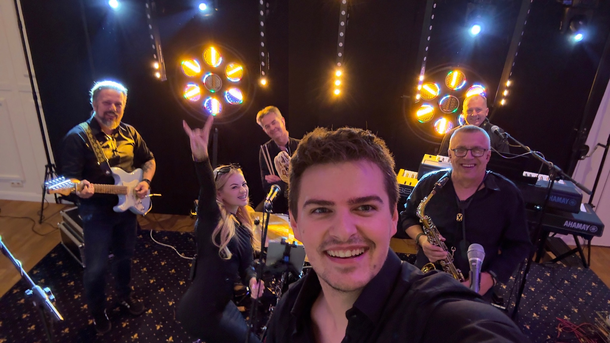 Zespół muzyczny w trakcie występu, widok z perspektywy pierwszego planu, uśmiechnięty wokalista, gitara, saksofon, instrumenty klawiszowe i oświetlenie sceniczne.