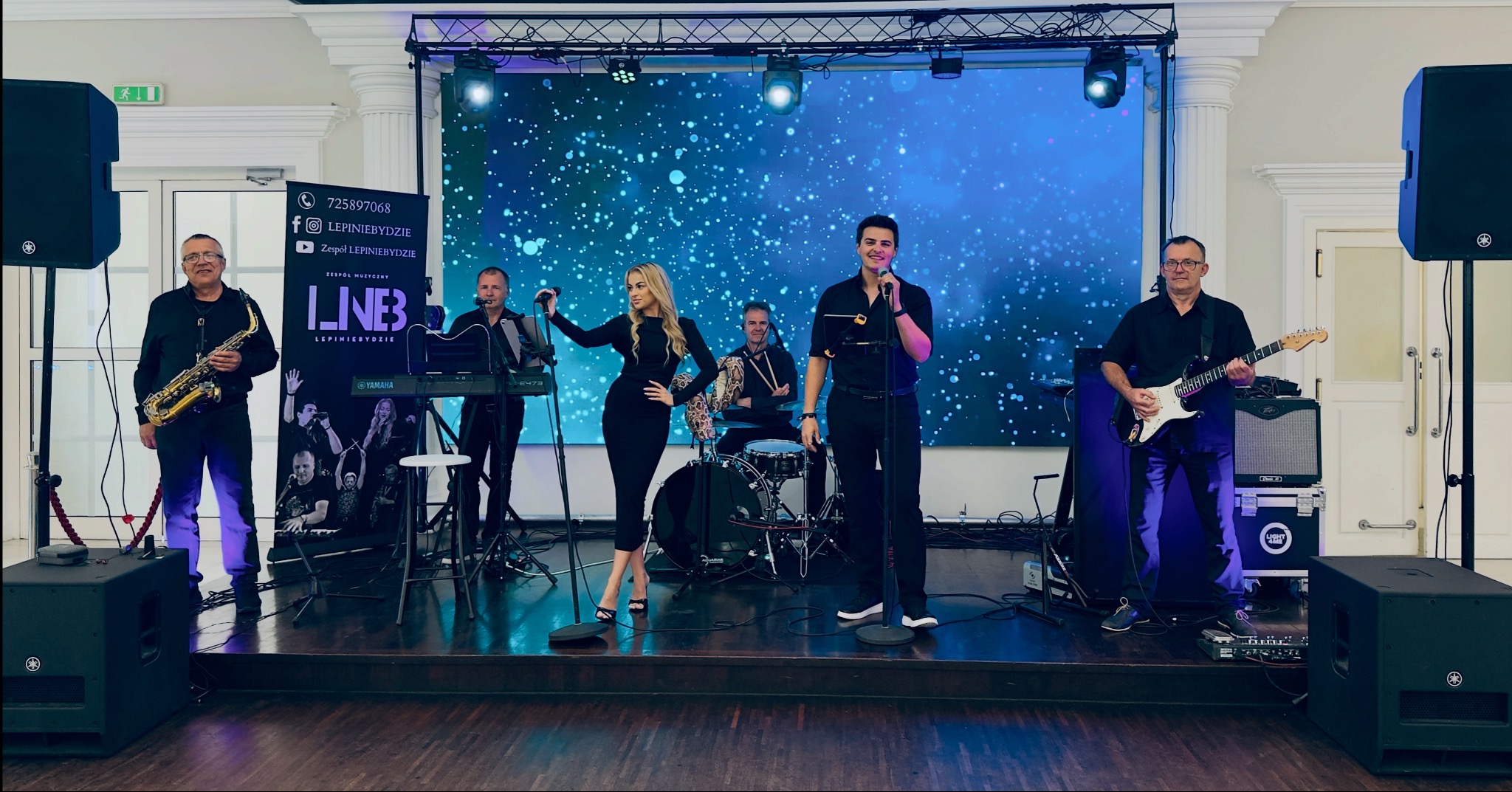 Zespół muzyczny na scenie z ekranem LED w tle. Wokalistka w czarnej sukience, saksofonista, gitarzysta, klawiszowiec i perkusista. Profesjonalne oświetlenie i nagłośnienie.