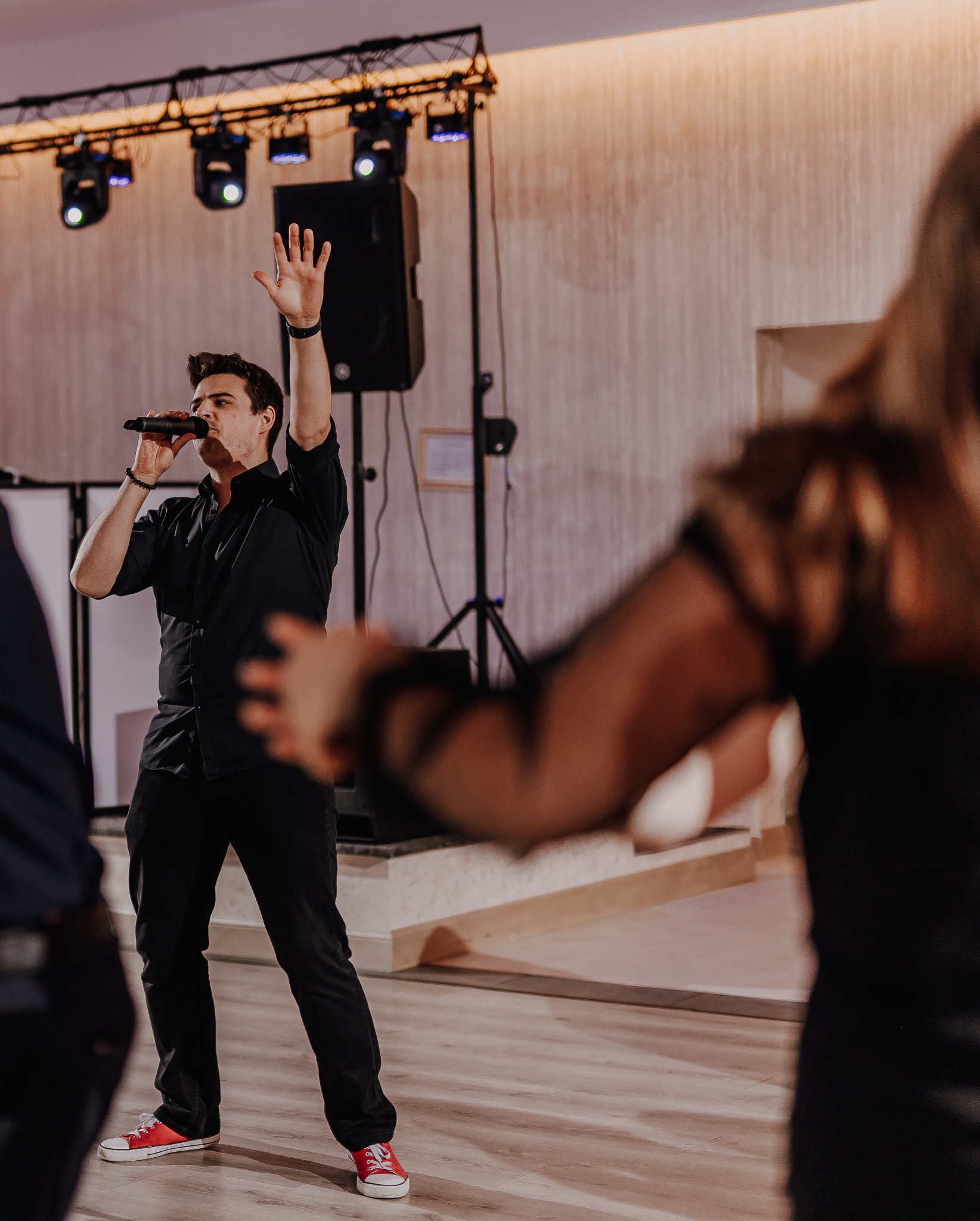 Wokalista w czarnym stroju i czerwonych trampkach śpiewa do mikrofonu na scenie, z podniesioną ręką, oświetlenie sceniczne w tle.